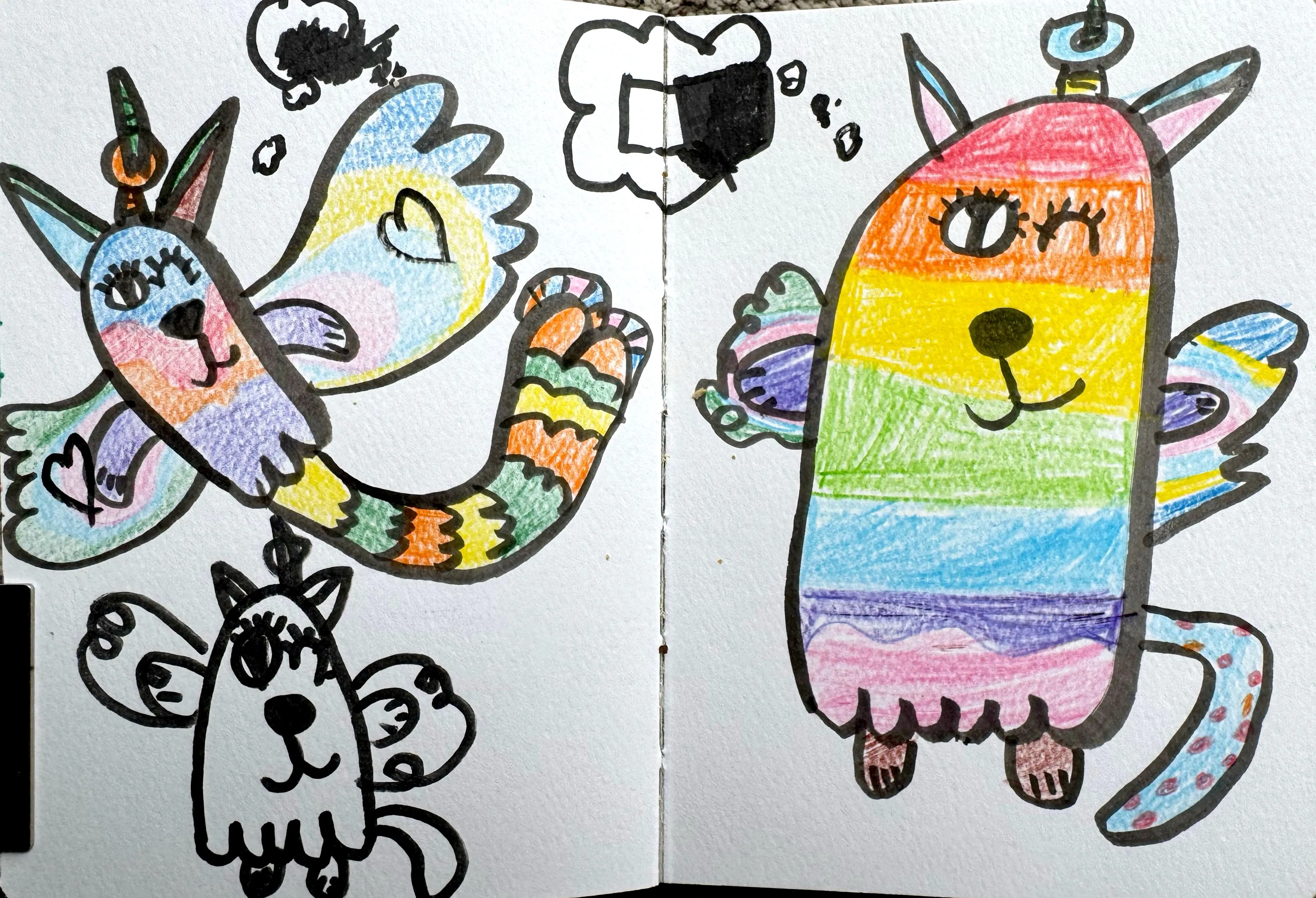 A sketchbook spread featuring several whimsical winged cat-unicorn creatures; one large figure coloured in pastel watercolour washes, one filled with bold rainbow crayon stripes, and one left as a black outline only.